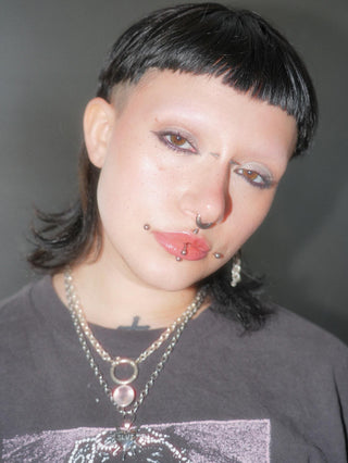 Person with black hair and piercings wearing a dark shirt against a gray background. They have on a thick sterling chain with a chunky rose quartz pendant hanging from it.